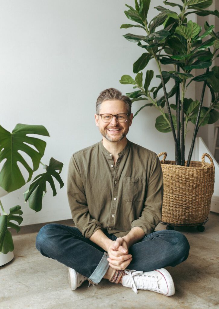 Jay Karr in an olive shirt, seated cross-legged on a concrete floor between two plants